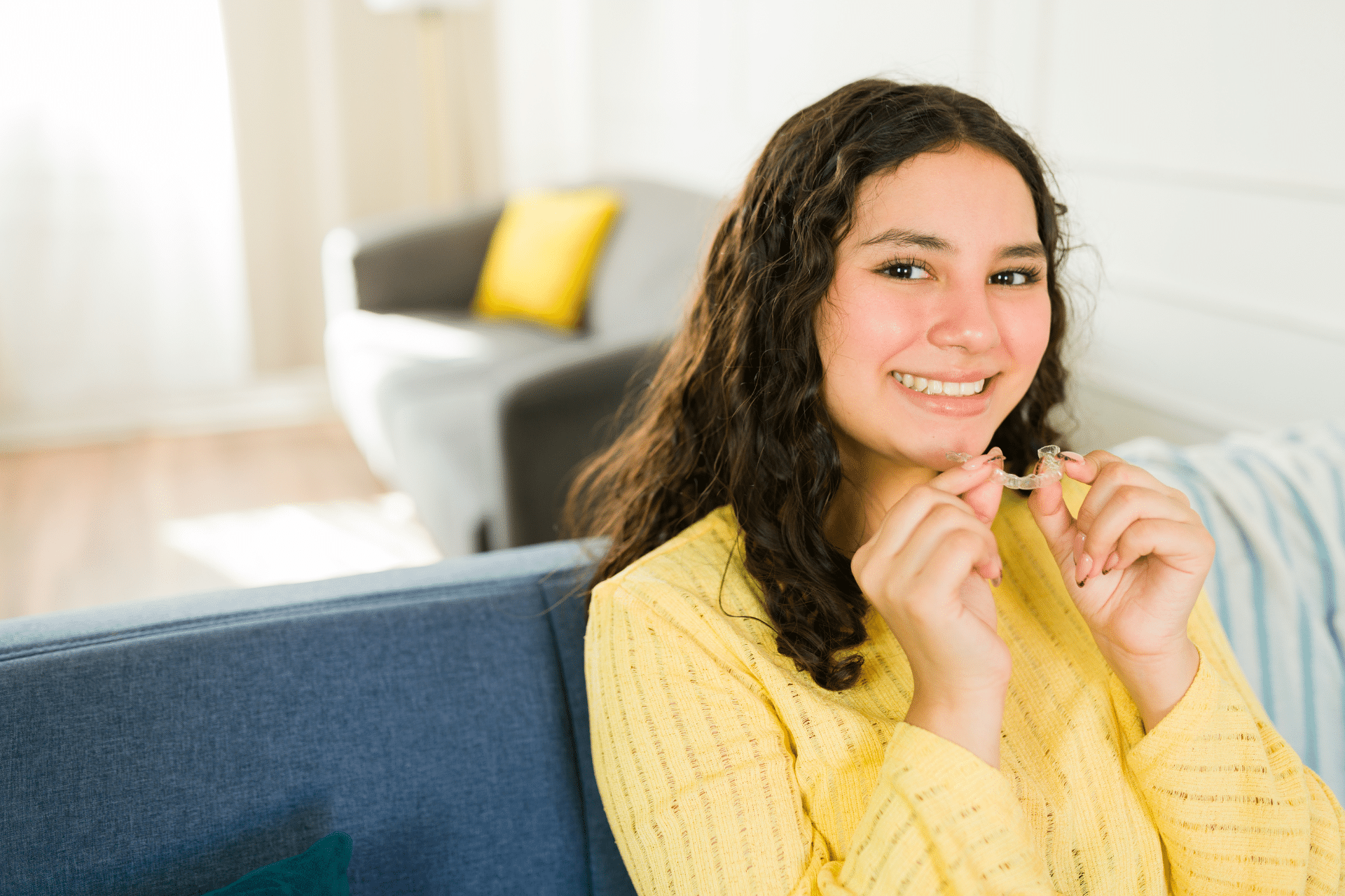Smiling teen holding a clear orthodontic aligner while sitting on a couch