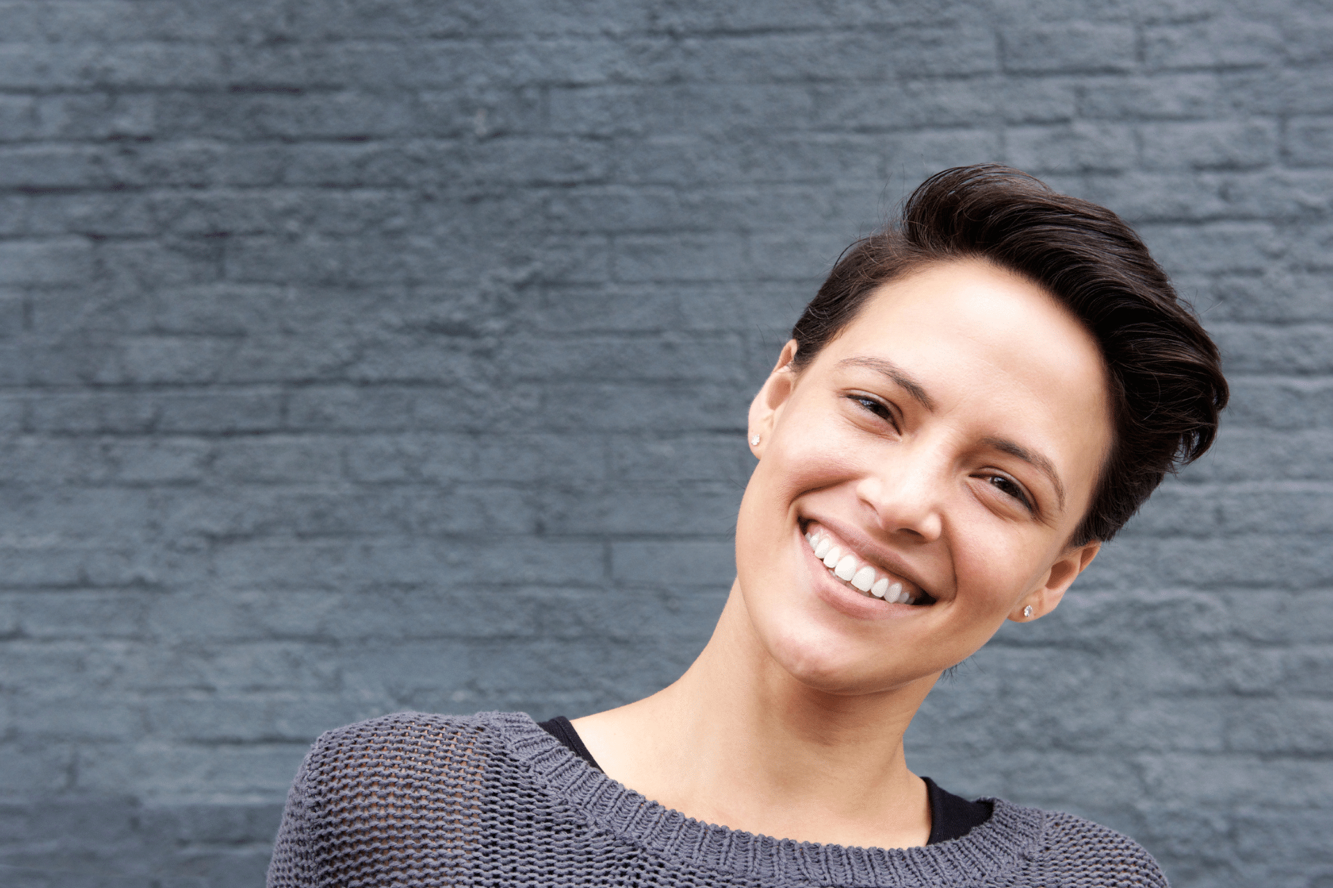 Smiling woman with short dark hair wearing a grey knit sweater in front of a dark brick wall